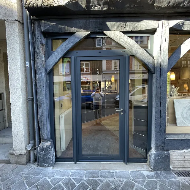 Une porte d'entrée moderne en aluminium gris anthracite intégrée dans une ancienne façade à colombages en bois.